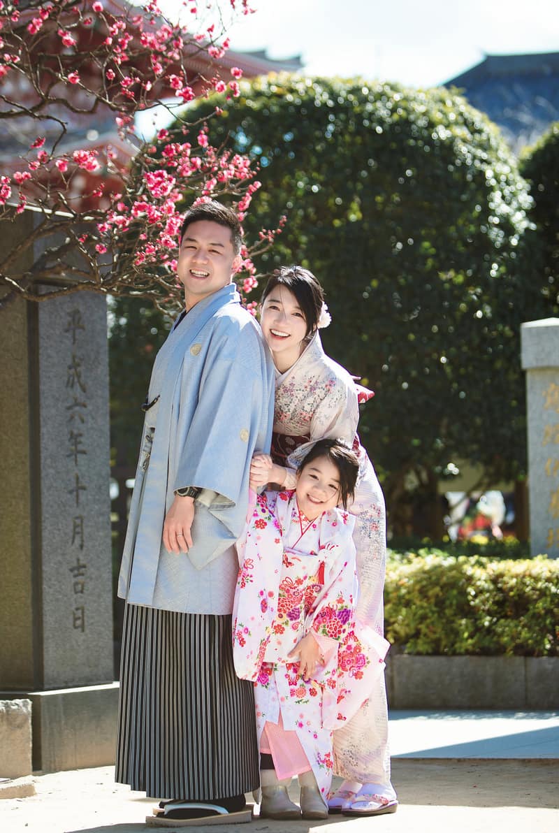紅梅の下で寄り添う三人。水色の羽織に縞袴の男性、白地に花の着物の女性、桃色の着物の女の子