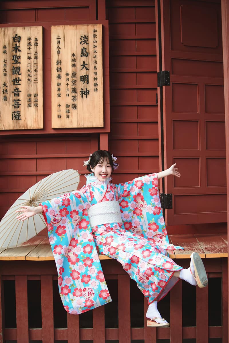 Guest in a turquoise cherry-blossom kimono with a sheer ruffled collar, holding a paper parasol on a wooden temple veranda