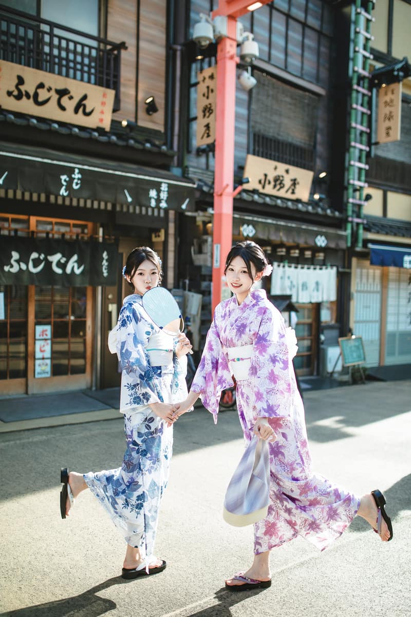 Two guests walking side by side along a street lined with shopfronts, one in a blue floral kimono holding a round fan, the other in a purple floral kimono