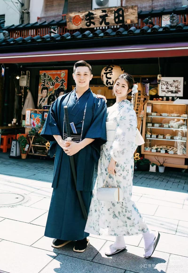 Two guests standing side by side under a shop awning, one in a deep blue men's kimono and one in a white kimono with a botanical print