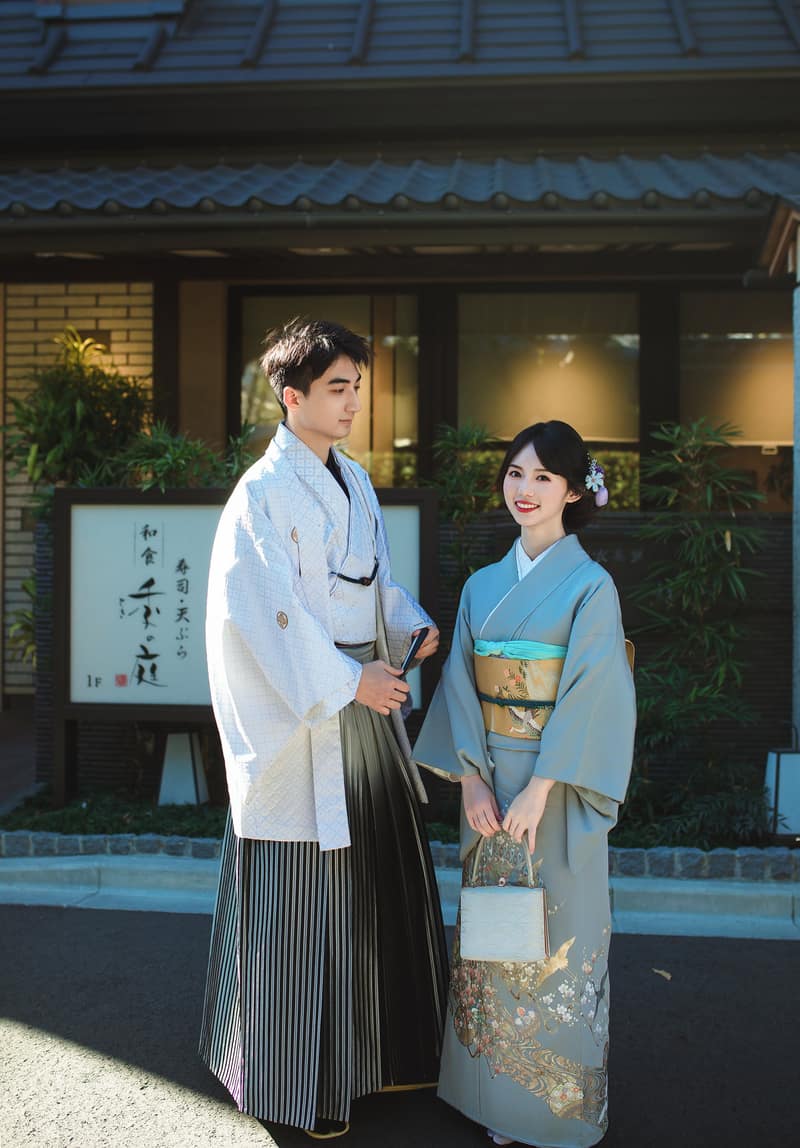 Two guests standing side by side in front of a tiled-roof building, one in a white haori over striped hakama, one in a pale blue kimono carrying a small bag