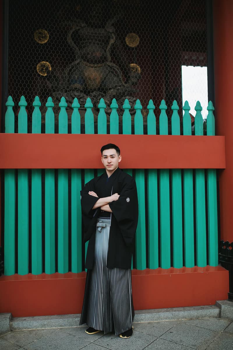 黒い羽織と銀灰色の縞袴姿で、朱と緑の社殿の格子の前に立つ男性のお客様