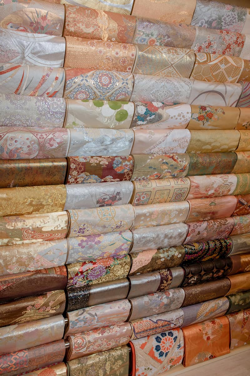 A wall of folded obi belts stacked on open shelves, gold weaves beside pastel patterns