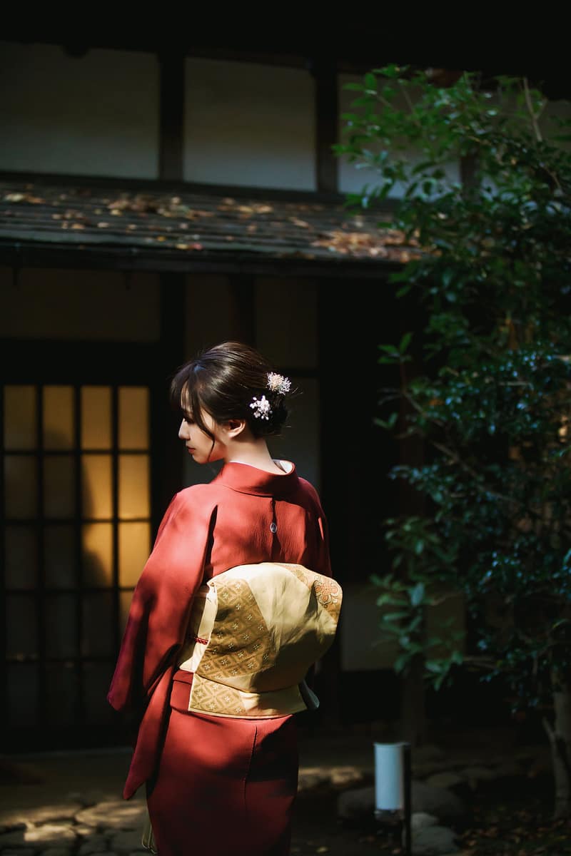 Back view of a vermilion solid-colour iromuji with a gold fukuro obi, in front of a wooden building