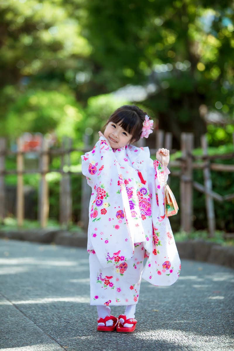 A child's kimono in white scattered with red and pink flowers, worn with a small drawstring pouch, standing on a paved path