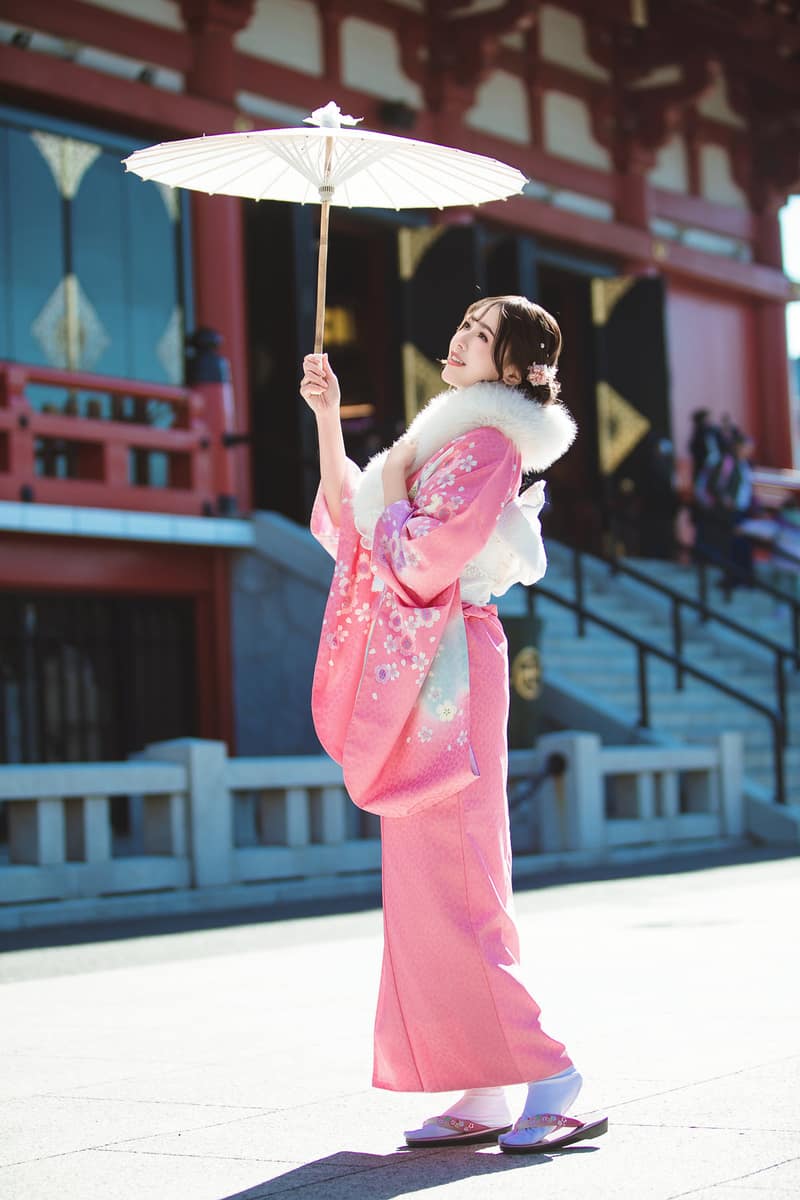 Pink short-sleeve furisode with cherry-blossom motifs, a white fur stole and a paper parasol, in front of a red temple hall